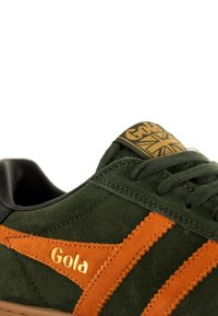 Sneakers in suede verde con strisce arancioni, dettagli neri e branding dorato. Presentano materiali testurizzati e dettagli cuciti.