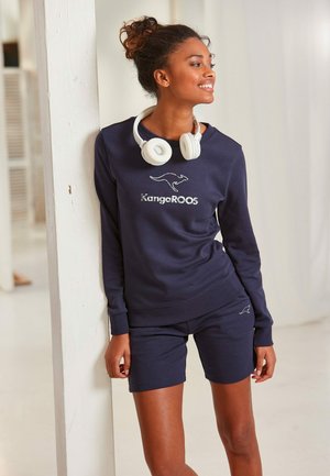 Navy sweatshirt and shorts set in soft fabric, featuring "KangeROOS" logo and kangaroo graphic, with white headphones.