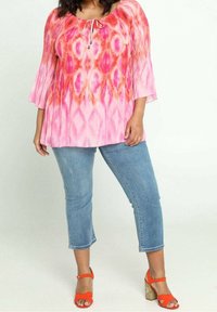 Donna che indossa una blouse a fantasia rosa e arancione, jeans blu a vita alta e sandali con tacco arancioni in piedi contro uno sfondo semplice.