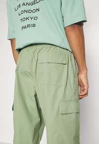 Ljusgröna cargo byxor med flera patchfickor, elastisk midja och en slät textur, parat med en mintgrön grafisk t-shirt.