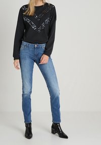 Nainen, jolla on yllään musta pitkähihainen paita, jossa on paljeteista tehtyjä chevron-kuvioita, siniset kapeat farkut ja mustat korkeat nilkkasaappaat, seisoo yksi käsi lähellä taskua.