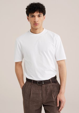 Młody mężczyzna z kręconymi włosami, noszący prostą białą koszulę z krótkim rękawem wpuszczoną w brązowe spodnie z wysokim stanem i plisowaniem, z plecionym paskiem.