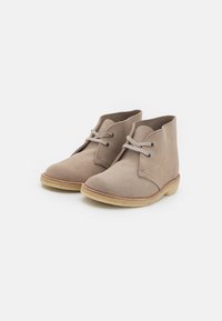 Beige suede ankle boots with rounded toe, lace-up front, and a light-colored rubber sole. Features soft texture and minimalistic design.