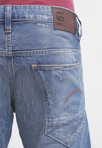 Jeans de mezclilla azul desgastados usados por una persona, mostrando el bolsillo trasero con costura naranja y una chapa de cuero de la marca en la cintura.