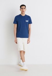 Tricou din bumbac de culoare albastru marin, cu logo alb "Lee", asortat cu pantaloni scurți crem și pantofi sport albi. Design simplu și confortabil.