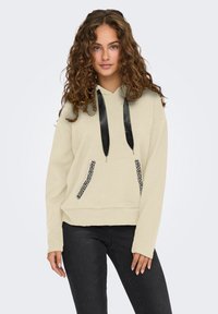 Giovane donna con capelli ricci che indossa un felpa beige con cappuccio e lacci in satin neri, con bordi delle tasche adornati, abbinata a jeans neri.