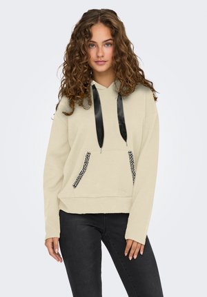 Giovane donna con capelli ricci che indossa un felpa beige con cappuccio e lacci in satin neri, con bordi delle tasche adornati, abbinata a jeans neri.