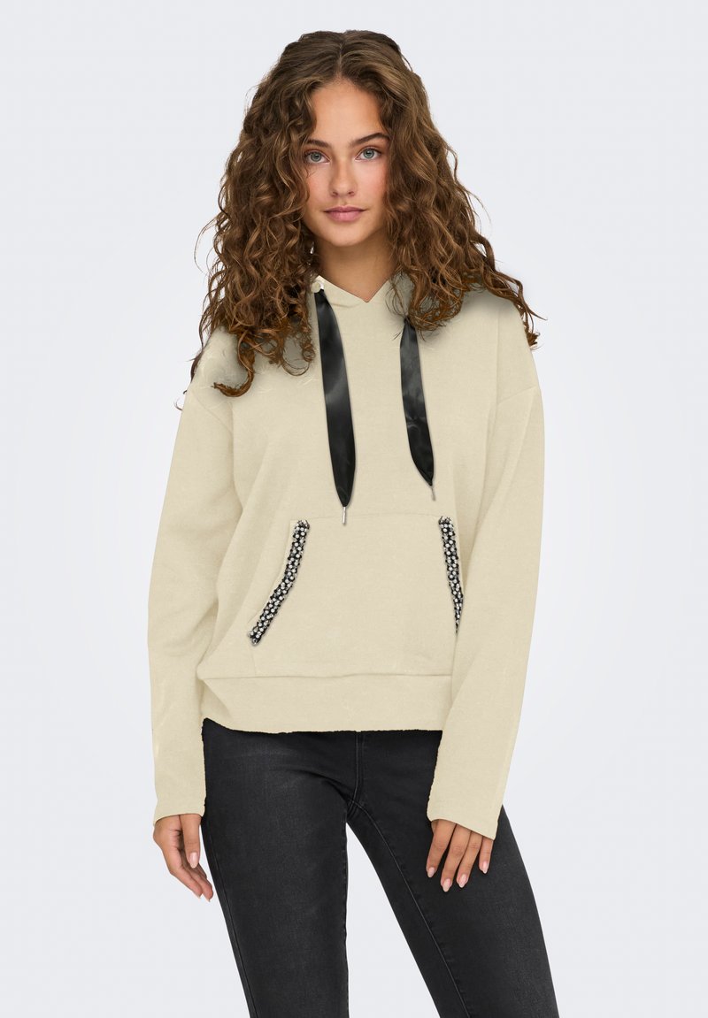 Giovane donna con capelli ricci che indossa un felpa beige con cappuccio e lacci in satin neri, con bordi delle tasche adornati, abbinata a jeans neri.