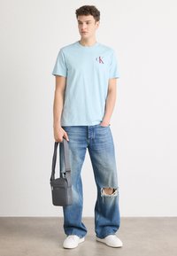 Svetle modré bavlněné tričko s malým burgundským logem na hrudi, v kombinaci se sepranými modrými džíny a šedou crossbody taškou. Bílé tenisky.