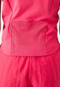 Camiseta deportiva rosa con un bolsillo trasero de malla transpirable. La tela es suave con un ligero brillo, complementada por unos pantalones cortos texturizados a juego.