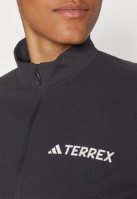 Giacca sportiva nera con colletto alto, chiusura a mezza zip e logo bianco "TERREX" sul davanti. Tessuto liscio e elasticizzato.