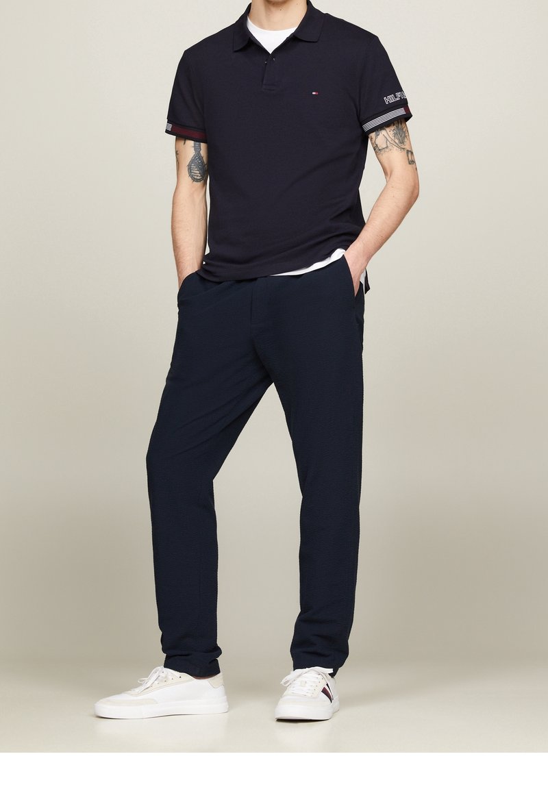 Homme portant un polo bleu marine foncé, pantalon assorti, baskets blanches, avec des tatouages sur les deux avant-bras, debout les mains dans les poches.