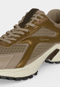 Baskets beige et or Calvin Klein avec tissu texturé, col matelassé, semelle épaisse et détail de la marque sur le côté et la languette.
