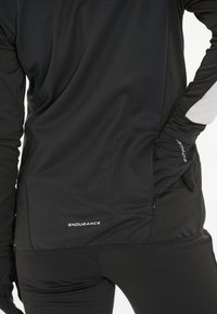 Schwarzes langärmliges Sportoberteil aus strukturiertem Stoff, mit einer Reißverschlusstasche, Daumenschlitzen in den Ärmeln und dem "ENDURANCE"-Logo am Saum.