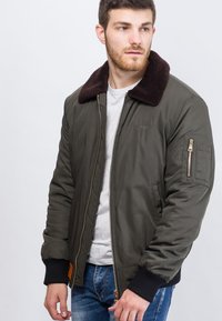 Olivgrüne Bomberjacke mit braunem Fellkragen, goldfarbenem Reißverschluss, gerippten Bündchen und einer mit Reißverschluss versehenen Armtasche; gepolsterter Innenraum sichtbar.