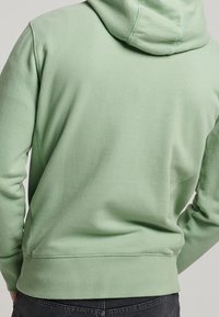 Grön luvtröja med mjuk textur, ribbstickade mudd- och midjeband. Har sömdekor längs ryggen och luvan.