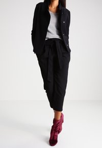 Giacca nera sopra una maglia grigia, abbinata a pantaloni neri a vita alta con cintura annodata e stivaletti in velluto color bordeaux.