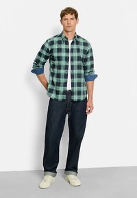 Grün-karierte und marineblaue Hemd mit Knöpfen, Ärmel hochgekrempelt, getragen über einem weißen T-Shirt, kombiniert mit dunklen Jeans und hellen Sneakern.