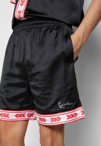 Shorts en satin noir avec un motif de flèche rouge et blanche au bas. Doté d'une taille élastique et d'un logo imprimé en blanc.