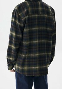 Camisa de franela en un patrón de cuadros verde, azul marino y crema. Cuenta con un cuello, mangas largas y costuras visibles en la parte posterior. Textura suave.