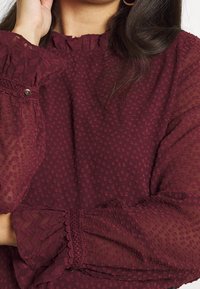 Burgunderfarbene Bluse aus strukturiertem transparentem Stoff, mit gerafftem Kragen und elastischen Bündchen, die Spitzenakzente und ein kleines Knopfdetail aufweisen.