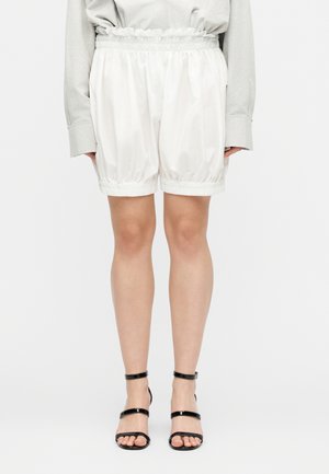 Fehér, bő szabású rövidnadrág rugalmas derék- és szegélypánttal, fekete pántos magassarkú szandállal és világos színű hosszú ujjú felsővel viselve.