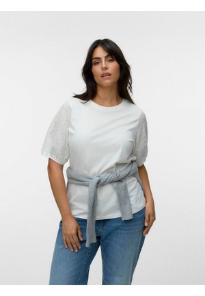 Kvinde iført hvid bluse med hulmønsterærmer, en grå sweater bundet omkring taljen og blå jeans, stående mod en ensfarvet lyse baggrund.