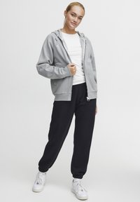 Light gray zip-up hoodie with adjustable drawstring, front pockets, paired with black tapered sweatpants and white sneakers. Smooth fabric texture.