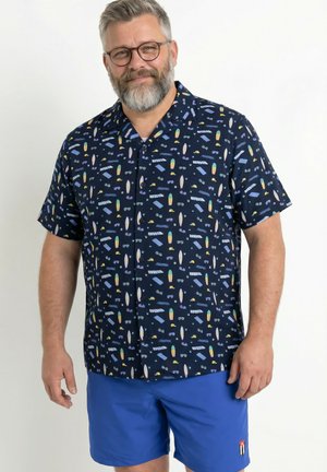 Mann mittleren Alters mit Brille und Bart, der ein marineblaues kurzärmeliges Hemd mit bunten Strandmotiven und leuchtend blaue Shorts trägt.