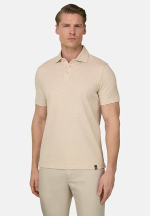 Homme portant un polo beige à manches courtes et un pantalon beige assorti, debout devant un fond clair uni.