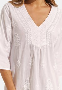 Witte tuniek met een V-hals, 3/4 mouwen en bloemenborduurwerk. Zachte stof met textuurdetails en geplooid ontwerp bij het lijfje.