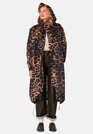 Vrouw die een capuchonjas met luipaardprint draagt over een beige trui, donkere opgerolde broek, witte sokken en zwarte schoenen, staand tegen een effen achtergrond.