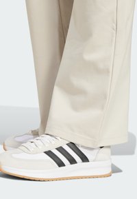 Adidas copalke v beli in svetlo sivi barvi s črnimi črtami; teksturiran usnje in mrežasti material; v kombinaciji z svetlo bež hlačami, vidnimi manšeti in gladkim materialom.