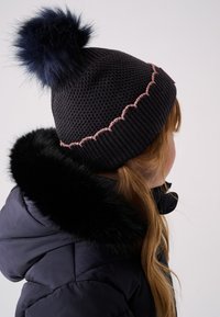 Neulottu musta pipo, jossa on laivastonsininen tekoturkistupsu ja vaaleanpunainen aaltoileva kuvio. Yhdistetty laivastonsiniseen toppatakkiin, jossa on turkisvuorinen kaulus.