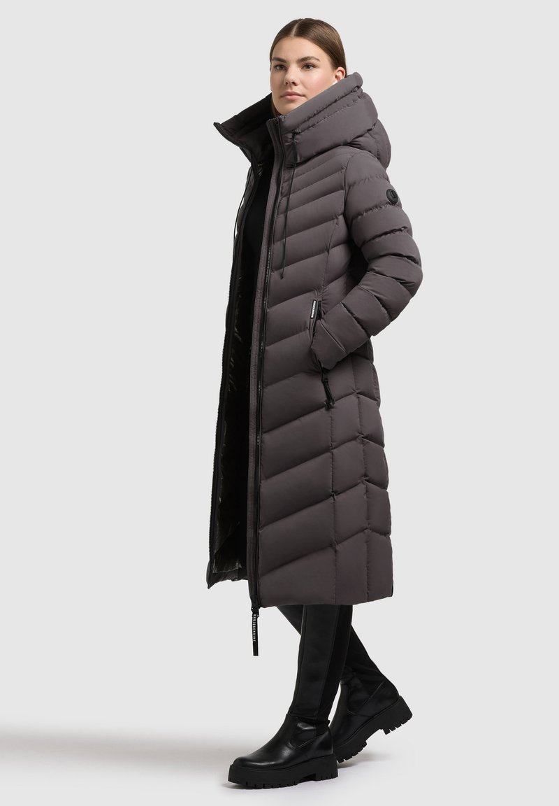 Eine Frau trägt einen langen, dunkelgrauen, gesteppten Daunenmantel mit einem hohen Kragen, Kapuze, seitlichen Taschen, schwarze Hosen und schwarze klobige Stiefel. Sie steht seitlich.