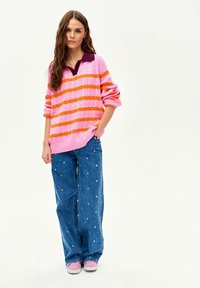 Kobieta z długimi włosami, ubrana w różowo-pomarańczowy sweter w paski, szerokie niebieskie dżinsy z małymi kolorowymi kropkami oraz różowe sportowe buty, stoi.