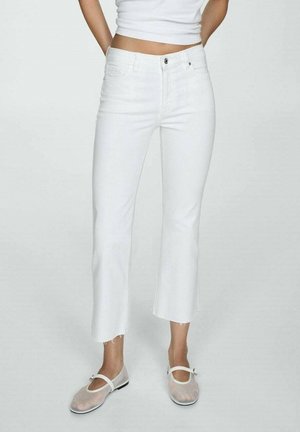 Person, die weiße Cropped-Jeans mit ausgefransten Säumen, ein weißes Cropped-Top und durchsichtige Mary Jane Ballerinas vor einem einfarbigen Hintergrund trägt.