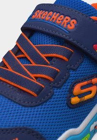 Μπλε παιδικό αθλητικό παπούτσι Skechers με πορτοκαλί λεπτομέρειες, κορδόνια σε ναυτικό μπλε με πορτοκαλί περίγραμμα και πολύχρωμο λογότυπο "S" στο πλάι.