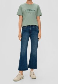 Grön bomullströja med "s.Oliver"-logotyp, tillsammans med mörkblå utsvängda jeans och beiga sneakers, som visar en avslappnad stil.