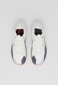Une paire de chaussures de sport blanches avec des accents noirs, orange et bleus, marquage Gore-Tex sur les côtés, vues d'en haut sur un fond blanc.