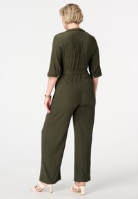 Combinaison vert olive avec des manches 3/4, taille ceinturée, tissu texturé et jambes larges ample. Portée avec des sandales à talons de couleur claire.