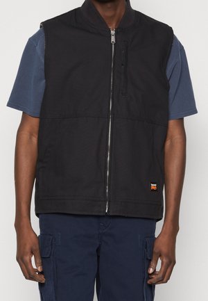 Homme portant un gilet noir zippé par-dessus une chemise bleue à manches courtes et un pantalon cargo bleu marine, debout avec les bras détendus le long du corps.