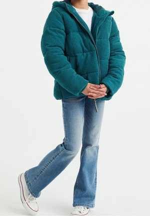 Barn iført blågrøn manchetvelour dunjakke, lyseblå udvidede jeans og hvide sneakers, står med hænderne foldet.