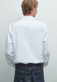 Weißes Langarmhemd mit einem Rückeneinsatz-Design, kombiniert mit dunklen Jeans aus Denim und einem geflochtenen schwarzen Gürtel. Texturiert und in lässigem Schnitt.