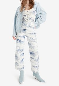 Chaqueta de mezclilla azul clara sobre una blusa blanca. Pantalones de talle alto con estampados de paisaje en azul. Botas azules de punta, a la altura del tobillo, con un pequeño tacón.