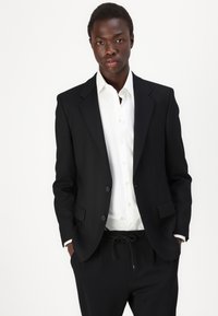 Blazer noir ajusté avec deux boutons et revers, porté sur une chemise blanche. Un pantalon noir à cordon complète l'ensemble.
