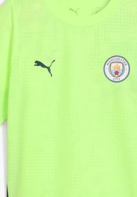 Maglia sportiva verde neon brillante con il logo Puma nero sul lato sinistro del petto e il badge del club di calcio Manchester City sul lato destro.