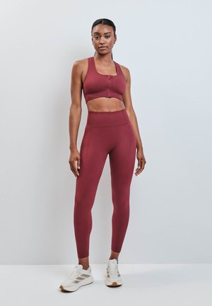 Vrouw staand, draag een bordeaux sportbeha met rits aan de voorkant en hoge leggings met witte sportschoenen tegen een effen achtergrond.