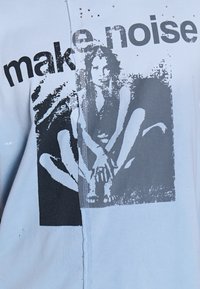 Μπλε πουκάμισο χωρισμένο με μαύρο σχέδιο μιας καθιστής γυναίκας και μερικώς ορατό κείμενο "make noise" στο στήθος.