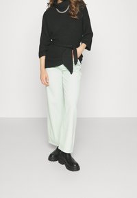 Suéter negro de punto con un lazo en la cintura, combinado con pantalones de pierna ancha verde claro y botas de tobillo negras gruesas. Collar de cadena de plata.